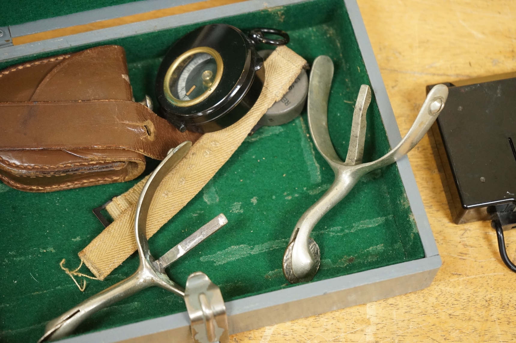 Three WWII military compasses including a Magnetic Marching Mark One compass, a small compass on a wrist strap and another in a leather case, together with a pair of steel spurs and a small grey painted wooden military b